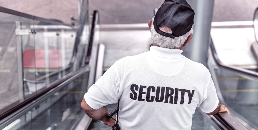 ein Security Mann mit einem weißen Shirt und Baseballmütze fährt eine Rolltreppe hinunter und wird von hinten fotografiert
